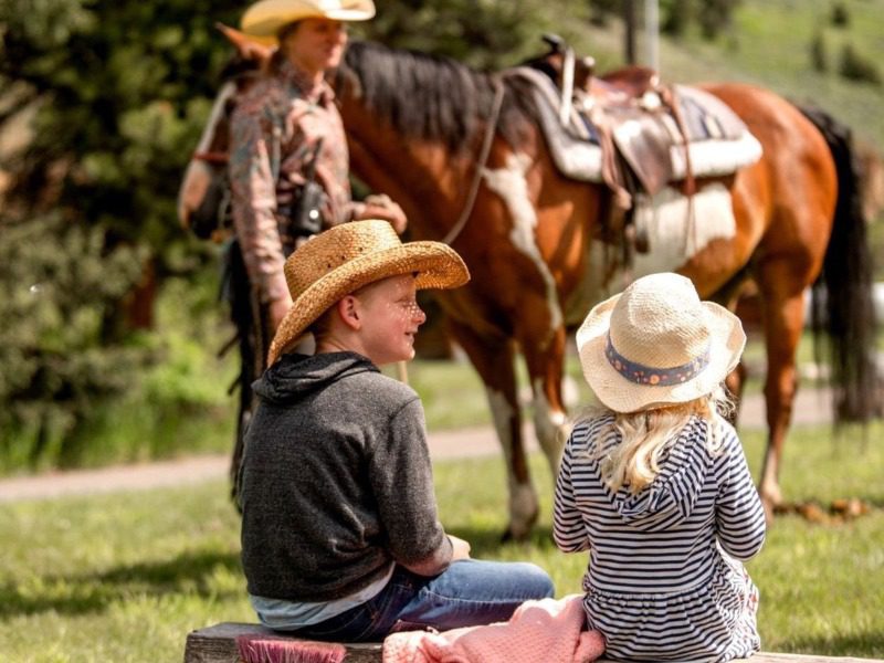 Montana Vacations for Families: A young brother and sister sit on a bench while watching horses and wranglers at work.